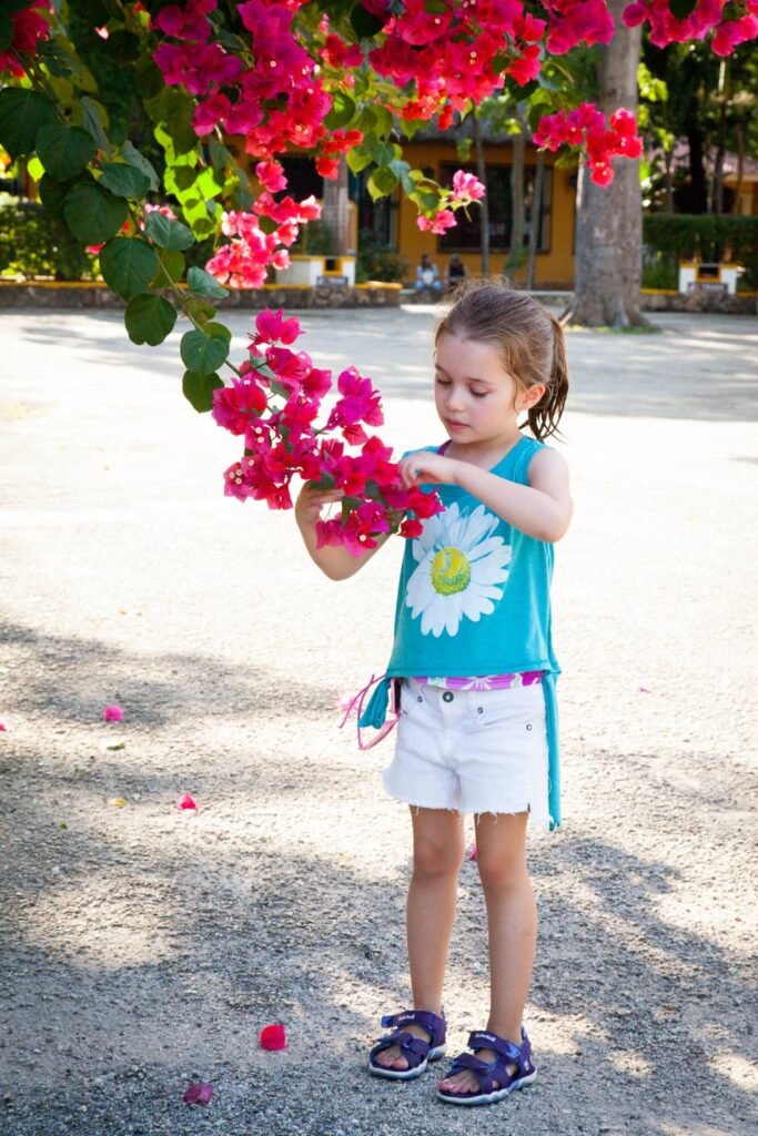A child stands beneath a lush bougainvillea blossom, wearing a daisy shirt and purple sandals.