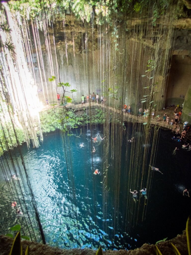 A deep cave sinkhole filled with bright blue water and covered in hanging vines. Swimmers enjoy the natural pool beneath the dense canopy.