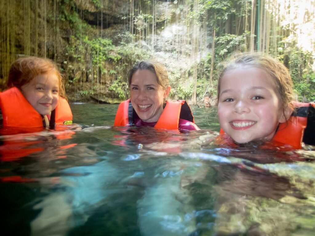 A group wearing orange life jackets smiles from a clear, green pool surrounded by lush vegetation.
