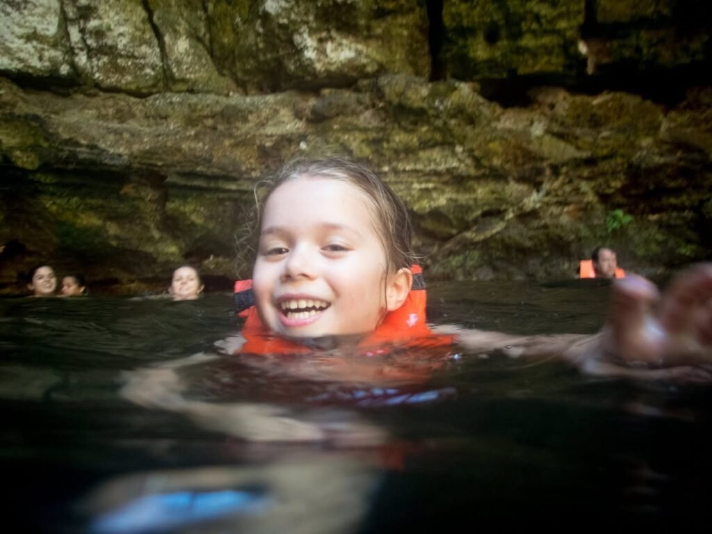 A child wearing an orange life jacket smiles while swimming in a natural river with rocky surroundings.