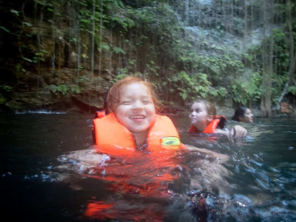 People wearing red life jackets swim in calm, dark water surrounded by dense tropical jungle foliage.