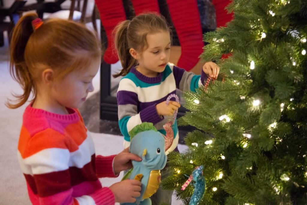 Two children in striped sweaters hang ornaments on a decorated Christmas tree. A dinosaur toy is held by one child during the festive activity.
