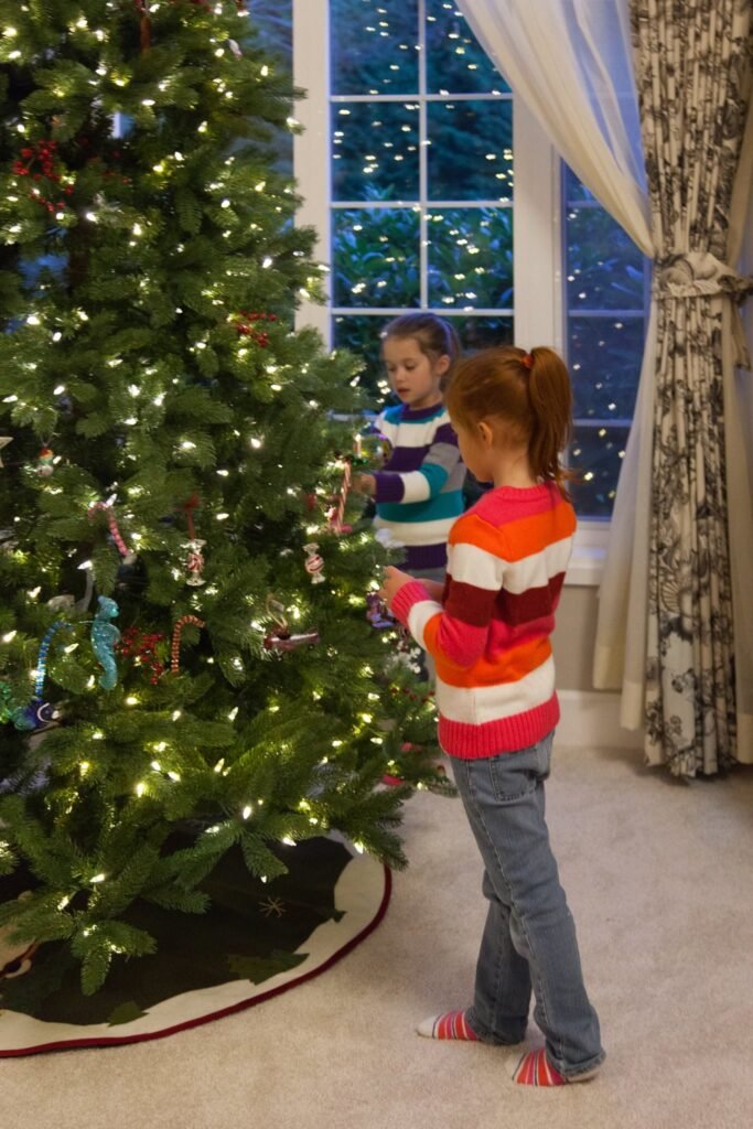 Two children placing ornaments on a lit Christmas tree near a large window.