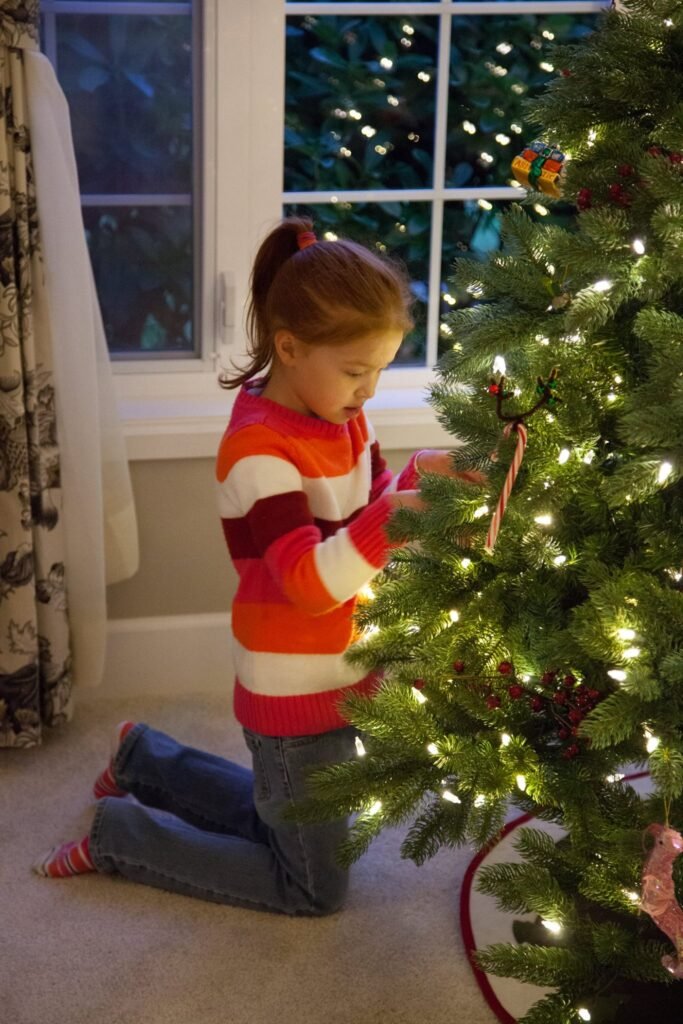 A child wearing a striped sweater kneels on the carpet to place an ornament on a lit Christmas tree.