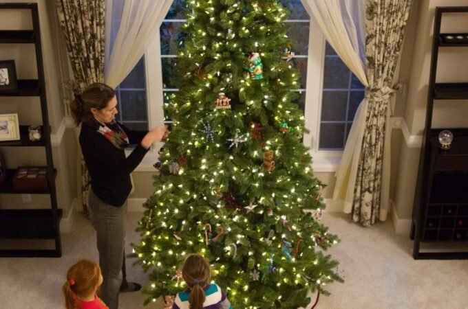 A family decorating a Christmas tree in front of a window with ornate curtains. Warm lights illuminate the tree in a cozy living room setting
