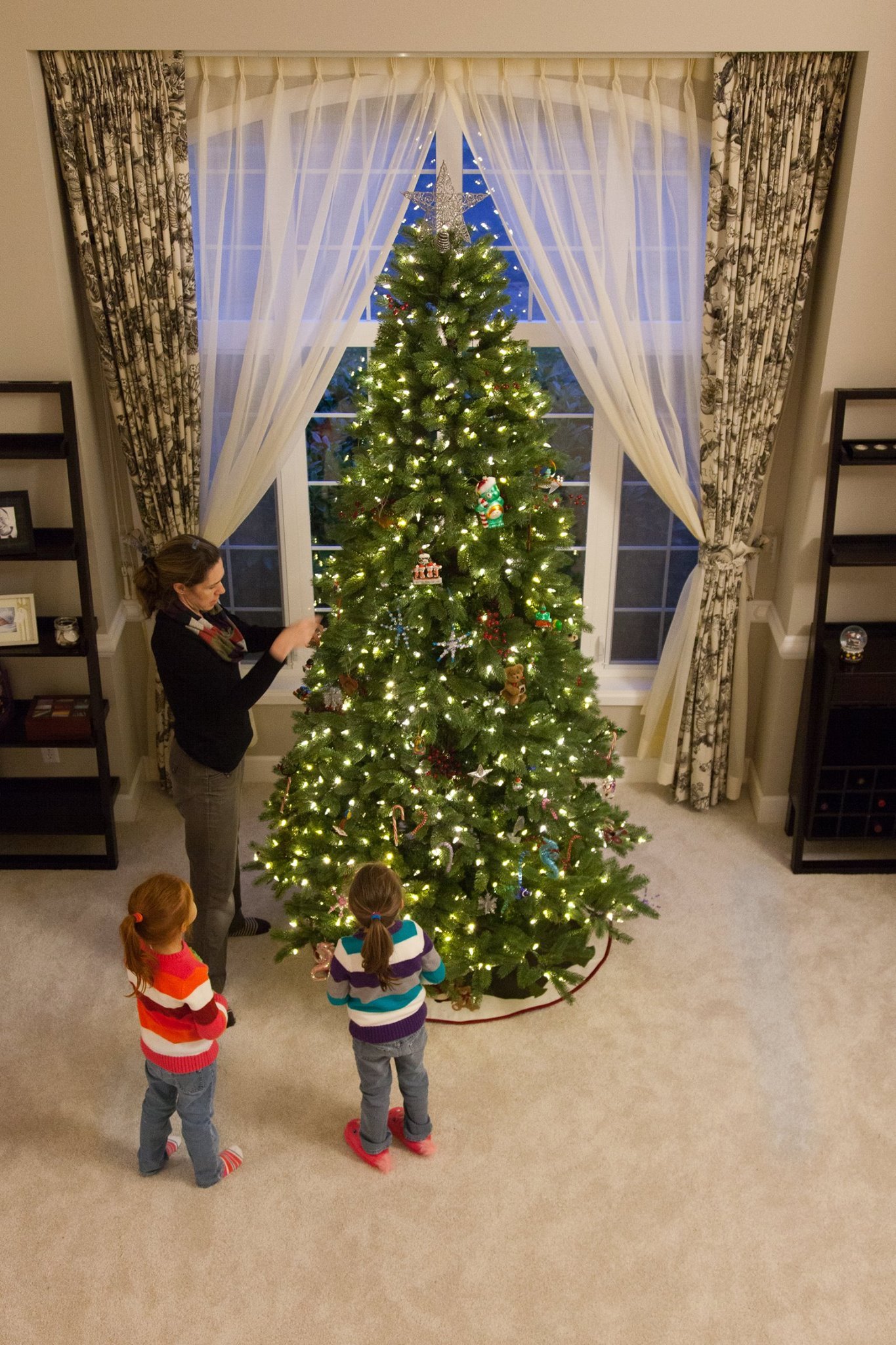 Trimming the Tree A family decorating a Christmas tree in front of a window with ornate curtains. Warm lights illuminate the tree in a cozy living room setting