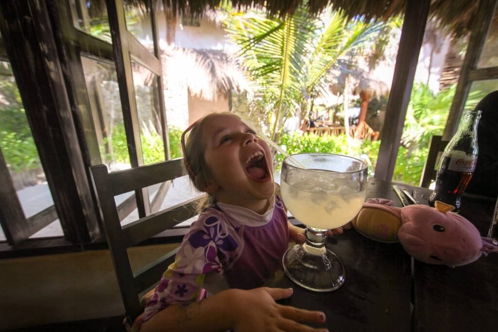 A laughing child sitting at a table with a drink in front of a window. Green foliage is visible outside.