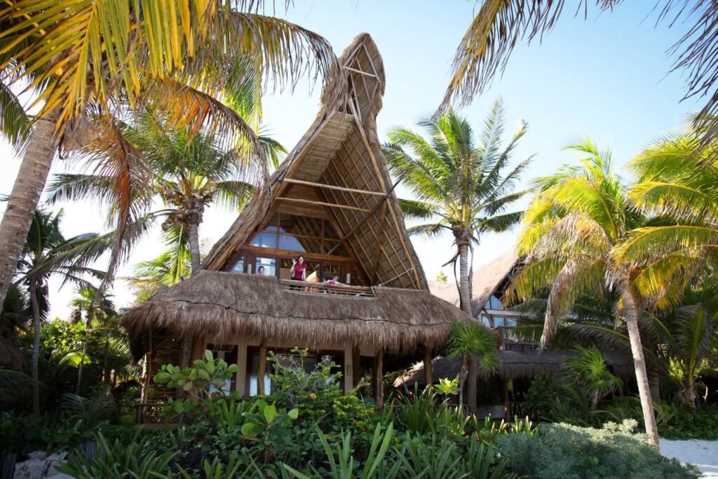 A thatched-roof bungalow surrounded by tall palm trees and dense tropical foliage. The structure blends naturally into the vibrant greenery of a tropical resort setting.