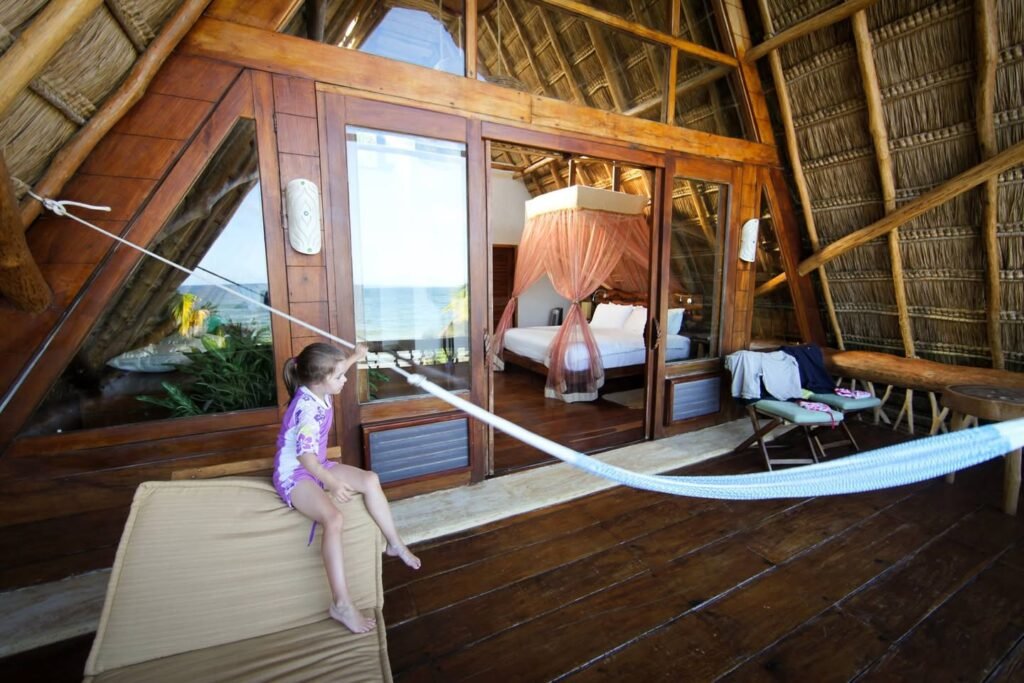 A person sitting on a cushioned bench inside a wooden and thatched treehouse, which features a hammock, large windows, and a four-poster bed.