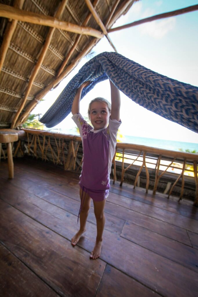 A child reaches up to touch a hammock under a thatched palapa roof by the ocean.