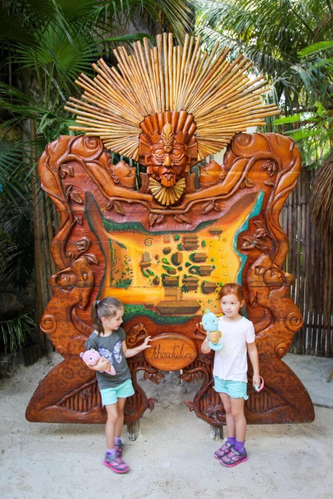 Two young girls stand in front of a large wooden sculpture resembling a map of the Rookery Bay ecosystem at a zoo or nature trail. The mural features a central feathered icon and tropical foliage in warm, earthy tones.