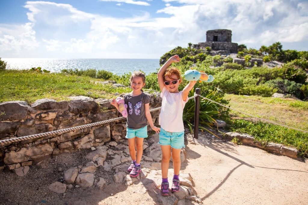 Two children standing near ancient ruins overlooking the ocean under a bright blue sky.