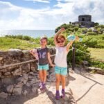 Two children standing near ancient ruins overlooking the ocean under a bright blue sky.