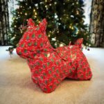 A gift wrapped in red festive paper shaped like a dog sits in front of a Christmas tree.