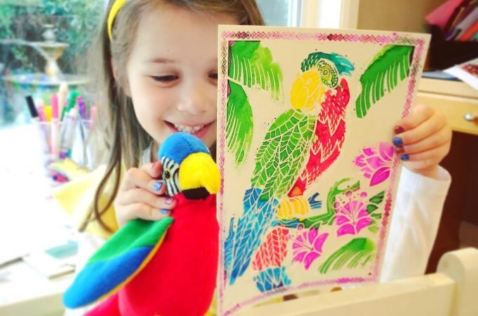 A young girl proudly displays a handmade parrot craft and a colorful patterned picture featuring tropical birds and flowers, standing indoors near a window by a table with supplies.