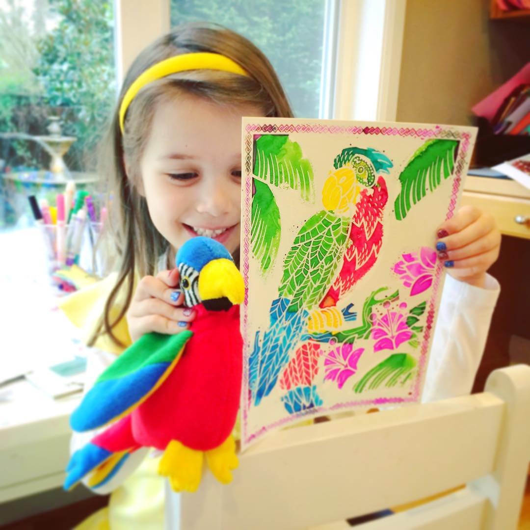 A young girl proudly displays a handmade parrot craft and a colorful patterned picture featuring tropical birds and flowers, standing indoors near a window by a table with supplies.