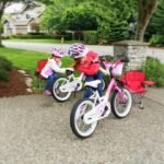 Children riding pink bicycles on a paved driveway with a lush green lawn and suburban house in the background.