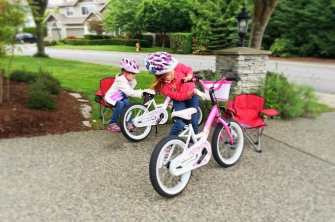 Children riding pink bicycles on a paved driveway with a lush green lawn and suburban house in the background.