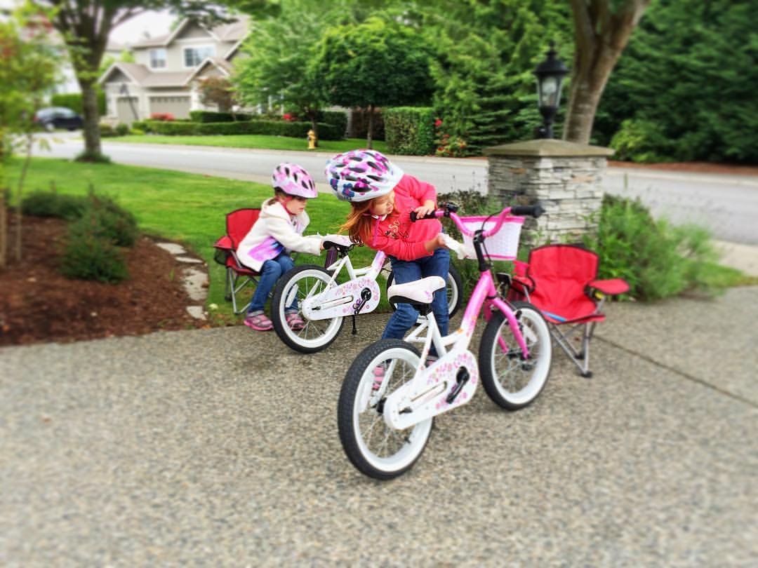 Children riding pink bicycles on a paved driveway with a lush green lawn and suburban house in the background.