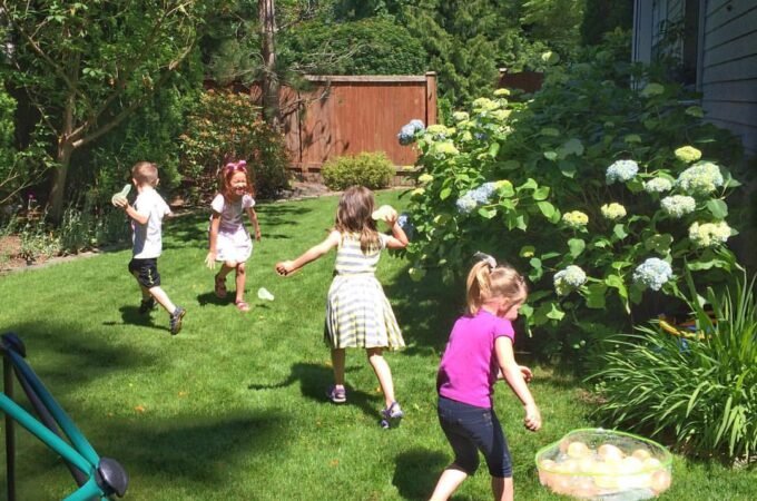 Children playing a beanbag toss game on a sunny lawn with a trampoline visible on the left.