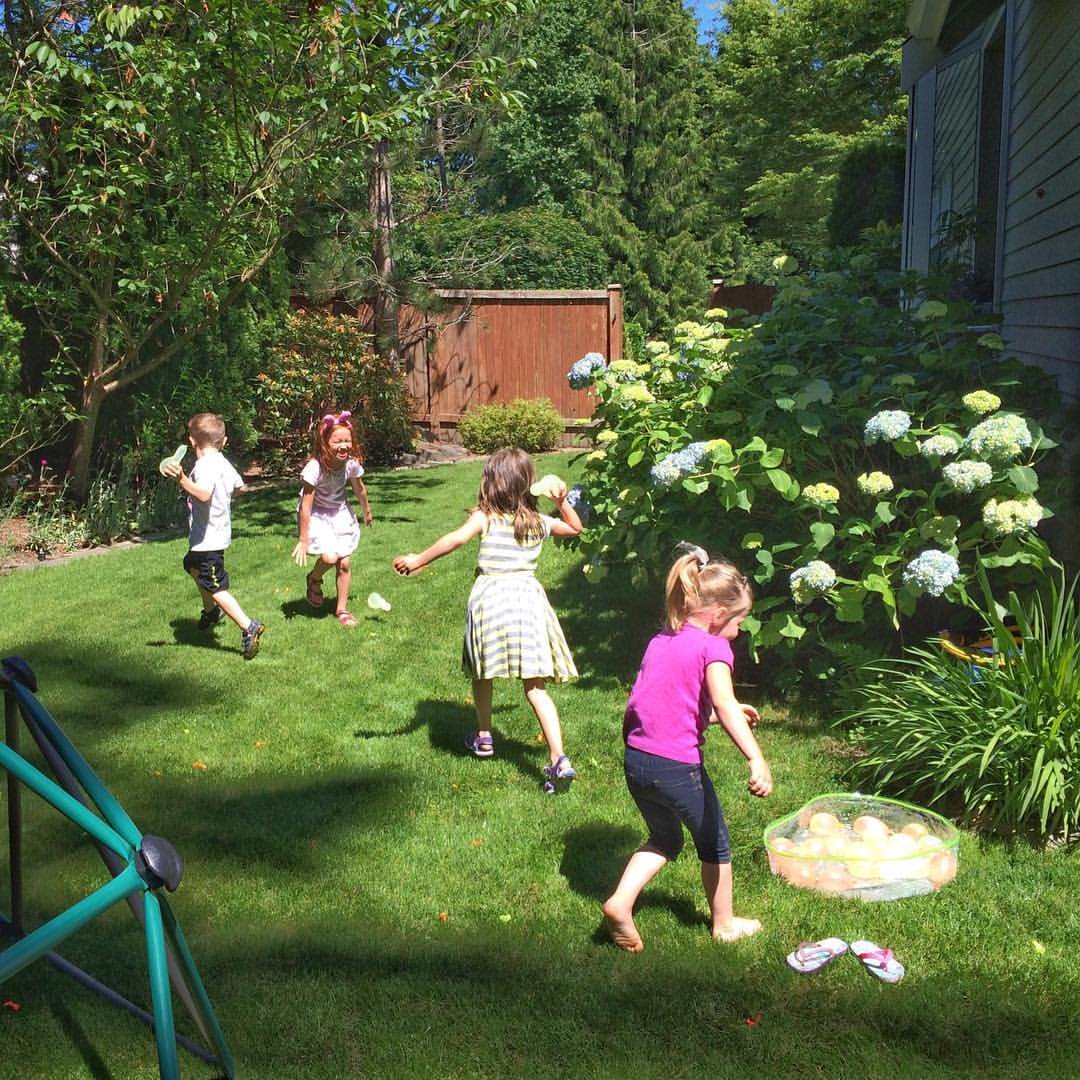 Water Balloon Fight Children playing a beanbag toss game on a sunny lawn with a trampoline visible on the left.