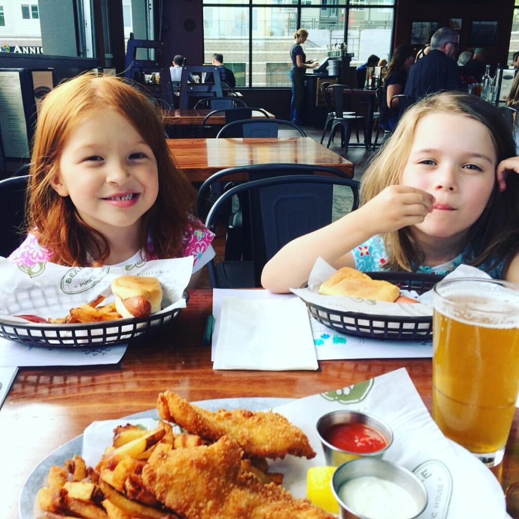 Two young friends share a meal of fish and chips with pint glasses in a sunny pub setting.