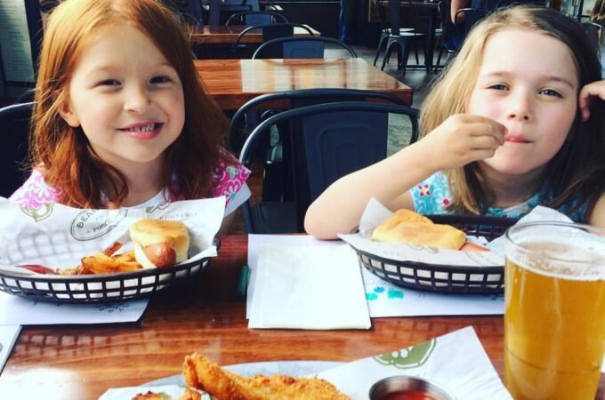 Two young friends share a meal of fish and chips with pint glasses in a sunny pub setting.