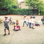 Children with helmets playing on a driveway with bikes, toys, and a car in the background on a sunny day.