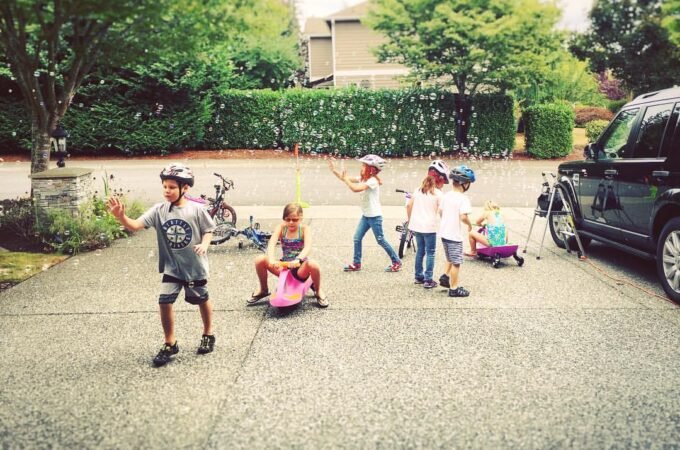 Children with helmets playing on a driveway with bikes, toys, and a car in the background on a sunny day.
