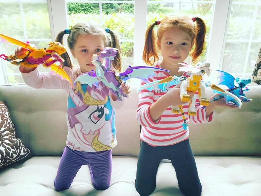 Two young children play with colorful LEGO Dragon toys on a couch in a bright, sunny room.
