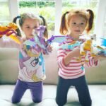 Two young children play with colorful LEGO Dragon toys on a couch in a bright, sunny room.