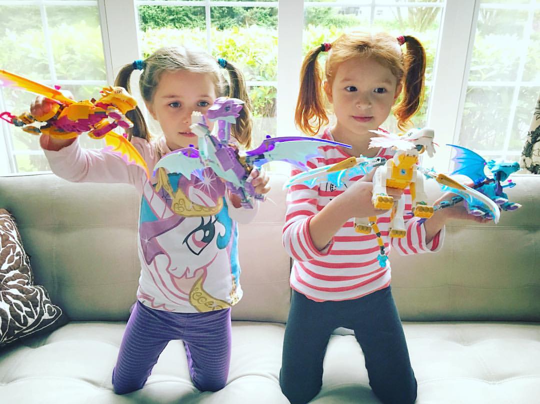 Two young children play with colorful LEGO Dragon toys on a couch in a bright, sunny room.