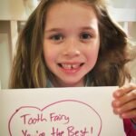 A smiling child holds a handmade sign reading 'Tooth Fairy You're the Best!' after losing their first baby tooth.