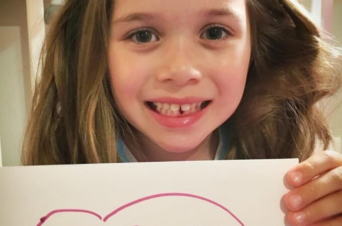A smiling child holds a handmade sign reading 'Tooth Fairy You're the Best!' after losing their first baby tooth.