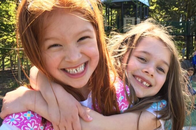 Two young girls share a sweet hug under bright sunlight in a wooded park, with a children's playhouse in the background.