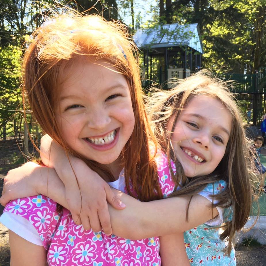 Two young girls share a sweet hug under bright sunlight in a wooded park, with a children's playhouse in the background.