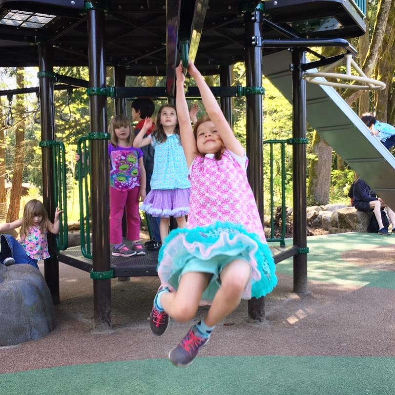 A child in a pink top and teal skirt hanging from a playground structure while friends look on.
