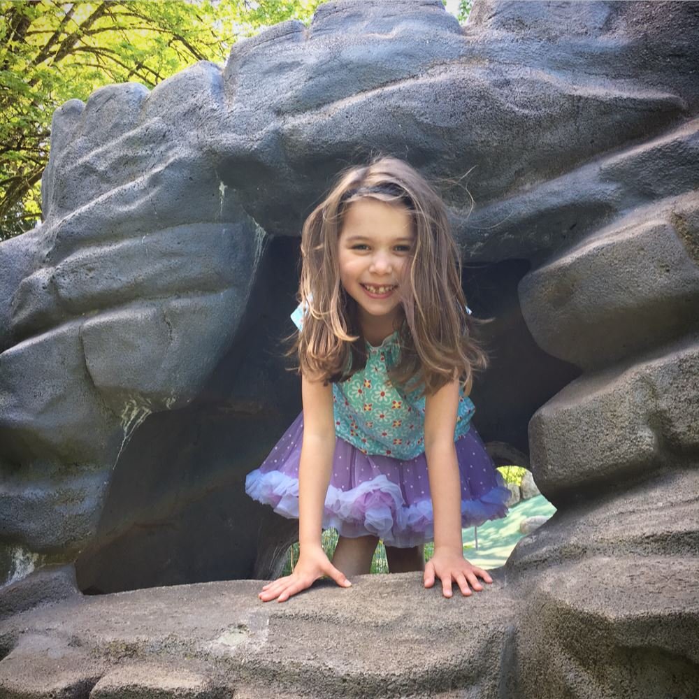 A smiling child poses in a rock grotto wearing a teal top and purple tutu skirt.