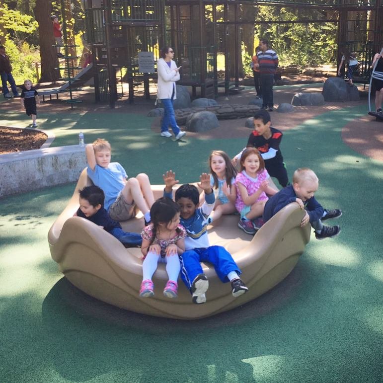 A group of young children sit together on a playground spinner in a sunny park setting.