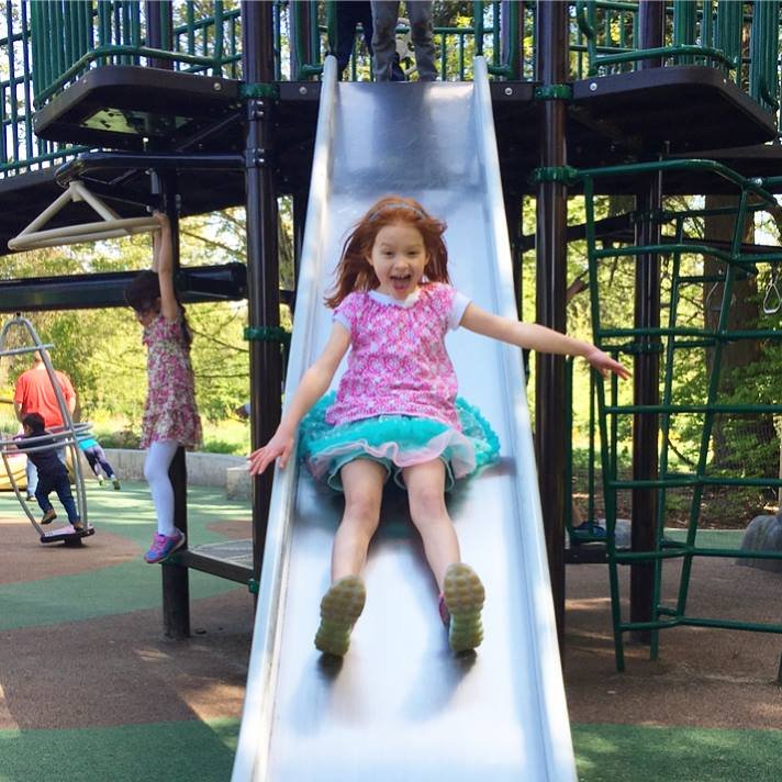 A child smiles while sliding down a playground slide on a sunny day. Other children are playing on the various playground equipment in the background.