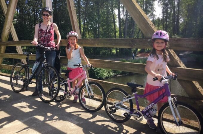 An adult and a child wearing helmets sit on bicycles on a wooden trestle bridge in a forest. A closer view of the adult at the handlebars is shown below.