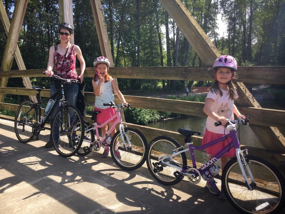 An adult and a child wearing helmets sit on bicycles on a wooden trestle bridge in a forest. A closer view of the adult at the handlebars is shown below.