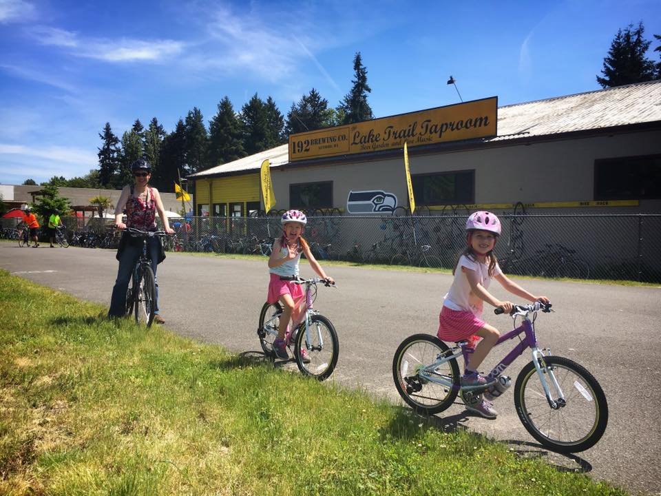 Children ride bicycles past a building labeled 'Bike Trail Supremacy' in a sunny outdoor setting. Tall evergreen trees and a clear blue sky frame the scene.
