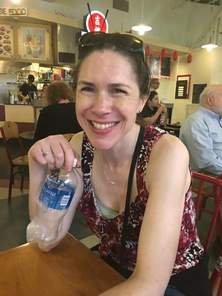 A person smiles while holding a water bottle at a table in a casual restaurant. The background shows a busy dining area with hanging decorations.