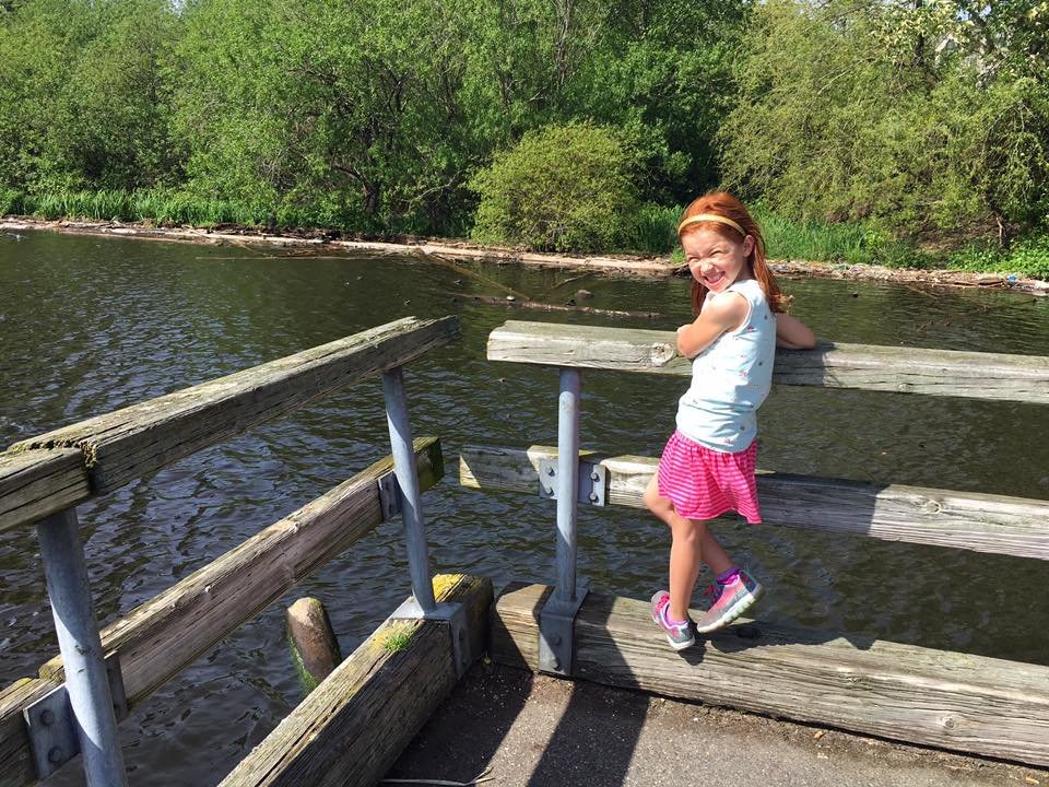A child stands on a wooden pier built over a calm river. Lush green trees and bushes line the water's edge in the background.