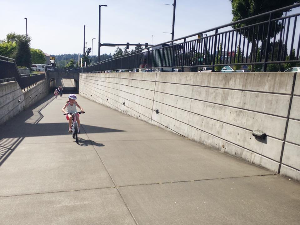 A person rides a bicycle along a concrete path beneath a bridge. The scene features railings and urban architecture.