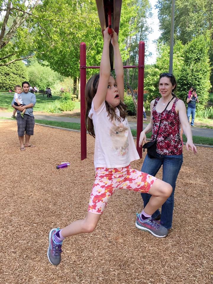 A child plays on playground equipment with an adult standing nearby. A sunny day at a local park.