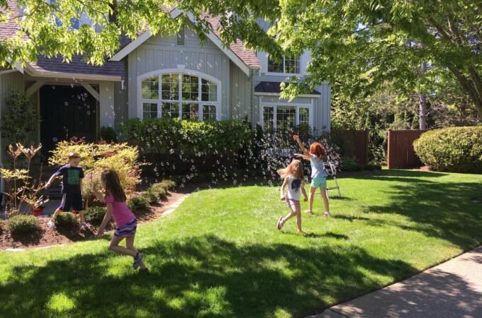 Children playing on a sunny front lawn surrounded by lush greenery before a suburban home.