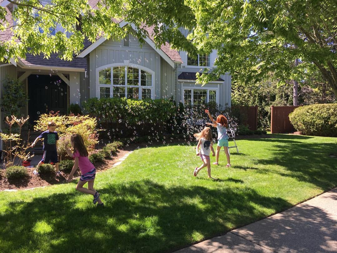 Children playing on a sunny front lawn surrounded by lush greenery before a suburban home.