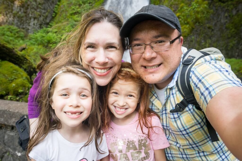 A family of four smiles for a selfie in a lush, green outdoor setting near a waterfall. The background features vibrant moss and natural stone textures.